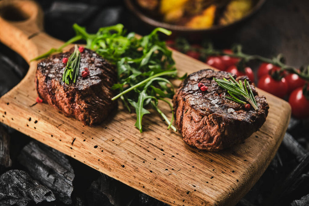 Grilled fillet steaks on wooden cutting board. Succulent thick juicy portions of grilled fillet steak served with tomatoes and roast potatoes on an old wooden board - Foto, imagen