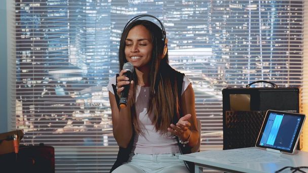Menina preta bonita em fone de ouvido cantando no microfone e gravando uma música em estúdio em casa
 - Foto, Imagem
