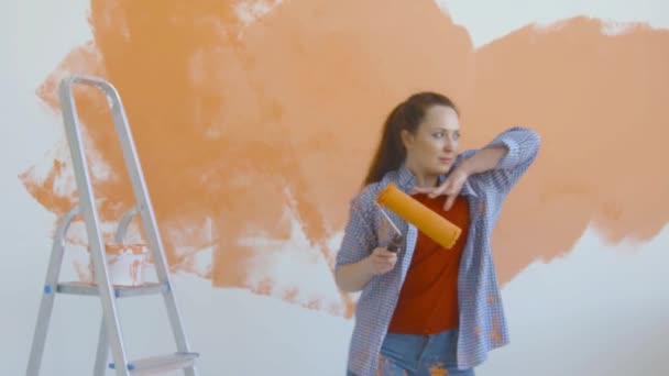 Singleton yaşam tarzı ve yenileme konsepti. Genç, gülümseyen bir kadın boya pateniyle dairedeki duvarı boyuyor ve dans ediyor. - Video, Çekim