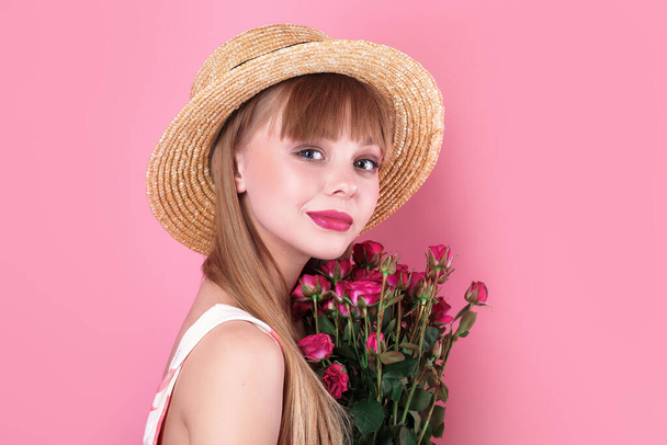 Belleza mujer rubia en sombrero de paja con flores frescas mirando a la cámara en el fondo del estudio rosa
. - Foto, Imagen