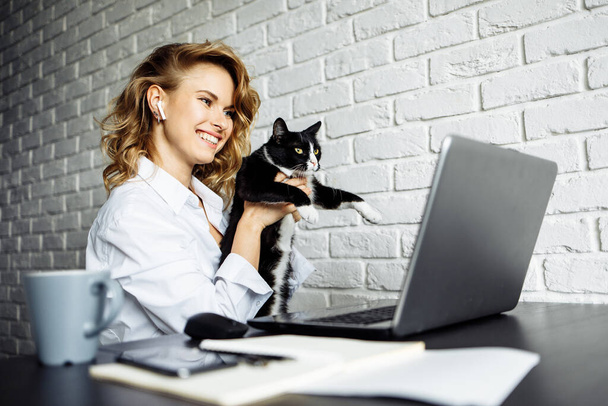 Young woman with black cat in her arms sitting at table with laptop - Фото, изображение