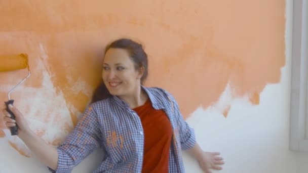 Singleton levensstijl en renovatie concept. Jonge lachende vrouw met schilderrol schildert de muur in het appartement en danst - Video