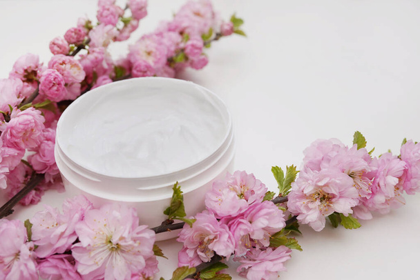 Geöffnetes weißes Glas mit natürlicher Kräutercreme für Frauen auf weißem Hintergrund mit blühenden rosa Blüten. - Foto, Bild