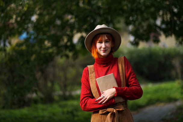 Portret młodej atrakcyjnej dziewczyny w czerwonych włosach w kapeluszu i czerwonym swetrze na tle zielonych drzew w parku. Trzyma książkę w rękach i czyta. - Zdjęcie, obraz