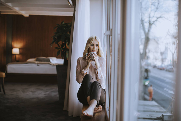 Mooie jonge vrouw met kopje thee of koffie zittend en drinkend op de vensterbank thuis - Foto, afbeelding