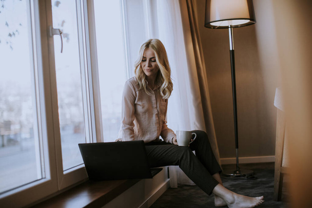 Hübsche junge Frau arbeitet am Laptop im Wohnzimmer auf der Fensterbank - Foto, Bild