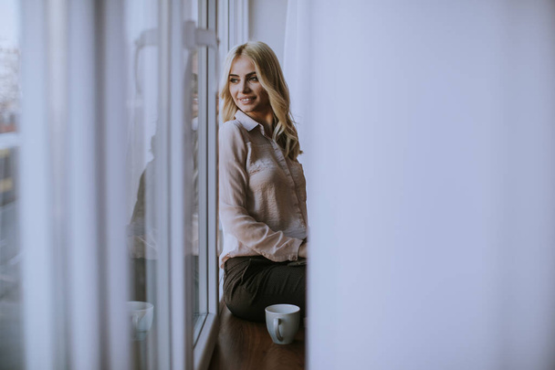 Kaunis nuori nainen kupin teetä tai kahvia istuu ja juo ikkunalaudalla kotona
 - Valokuva, kuva
