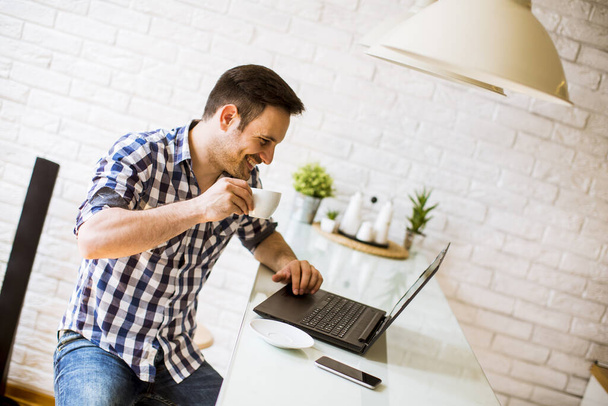 Beau jeune homme assis sur le bureau de la cuisine à la maison et en utilisant un ordinateur portable et en buvant du café
 - Photo, image