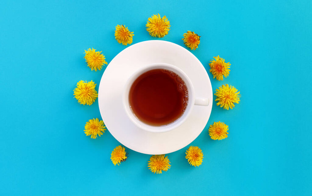 Cup of herbal tea over blue paper  background decorated with yellow dandelions flowers and green leaves. 5 o'clock concept. Flat lay, top view, copy space. - Фото, изображение