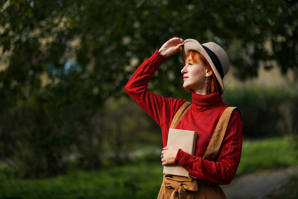 Headshot portrait of a young attractive girl with red hair in hat and red sweater on background of green trees in park. She holds book in hands and reads. - 写真・画像