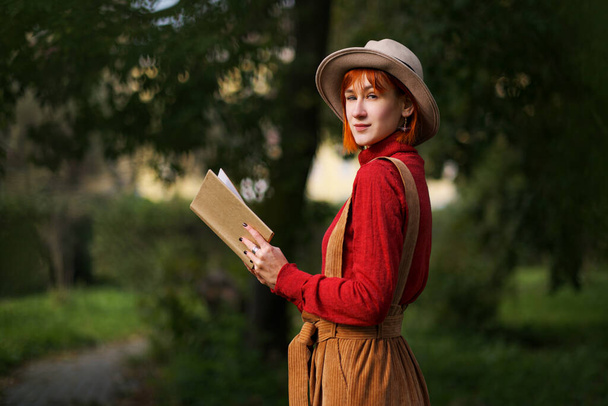 Portret młodej atrakcyjnej dziewczyny w czerwonych włosach w kapeluszu i czerwonym swetrze na tle zielonych drzew w parku. Trzyma książkę w rękach i czyta. - Zdjęcie, obraz