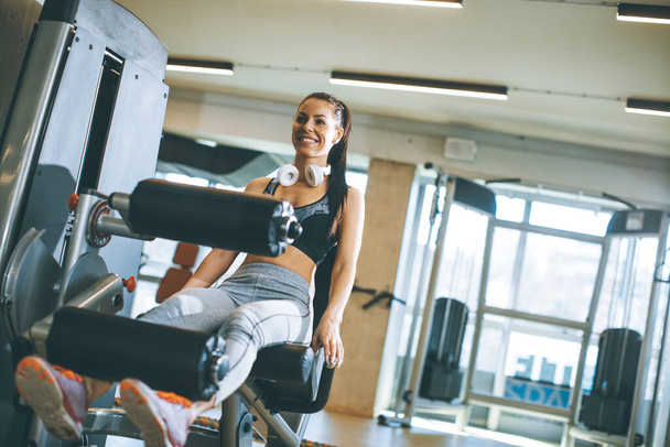 Pretty young woman having exercises on leg extension and leg curl machine in the gym - Foto, Imagem
