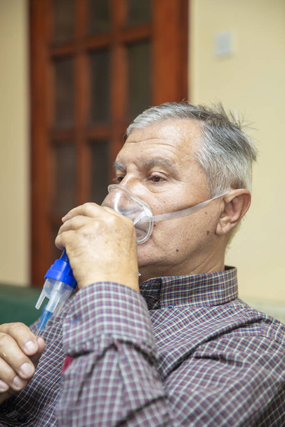Ανώτερος άνθρωπος που χρησιμοποιεί ιατρικό εξοπλισμό για εισπνοή με αναπνευστική μάσκα, νεφελοποιητής στο δωμάτιο - Φωτογραφία, εικόνα