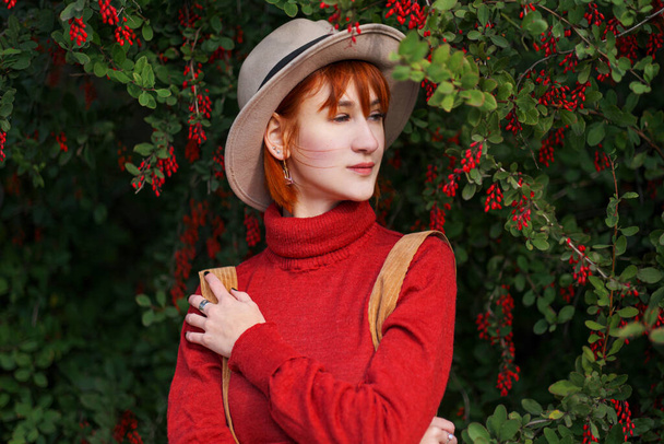 Portrait of a young attractive girl with red hair in a hat and a red sweater against the background of a bush of barberry with red berries. - Foto, imagen
