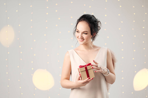 Young brunette woman in evening dress on a white background. Holding Christmas present box. Joy, happiness and surprise. - Фото, изображение