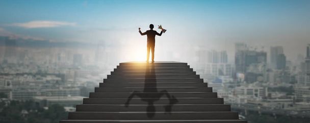 Концепция достижения высшей цели и успеха, успешный бизнесмен, ведущий бизнесмен, победитель, взбирающийся на вершину высокой лестницы города показывает руку вверх, держа награду, трофей инвестиционного шага
 - Фото, изображение