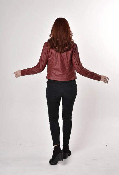 Portrét hezké dívky s rudými vlasy v černých džínách a botách s koženou bundou. celková délka stojící póza s zpět do kamery studio pozadí. - Fotografie, Obrázek