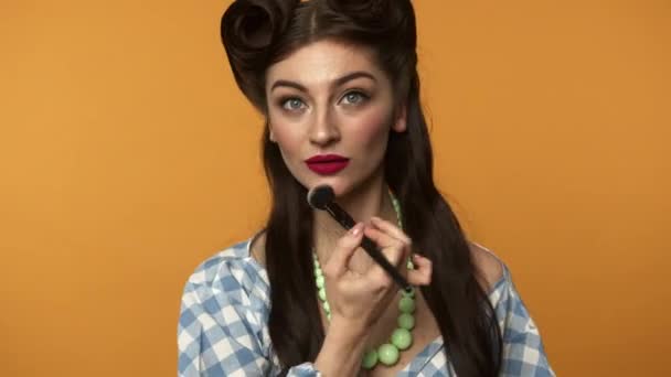 Cheerful pin up woman with brush applying powder on face and winking at camera - Footage, Video