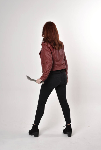 Портрет красивой девушки с рыжими волосами в черных джинсах и сапогах с кожаной курткой. полная длина стоящая поза держа нож студийный фон
. - Фото, изображение