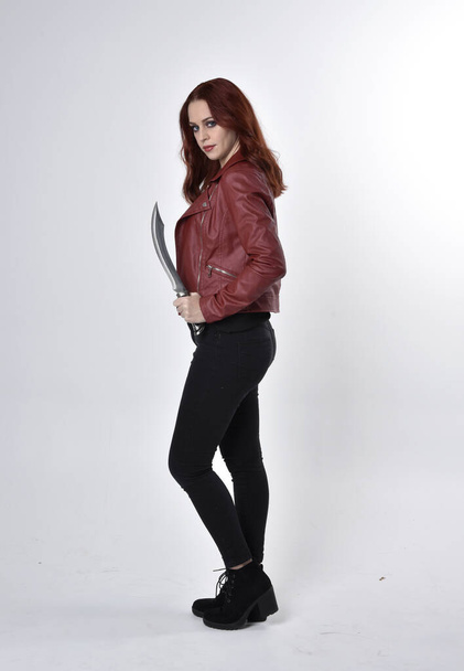 Portrait of a pretty girl with red hair wearing black jeans and boots with leather jacket.  full length standing pose holding knife a studio background. - Photo, Image
