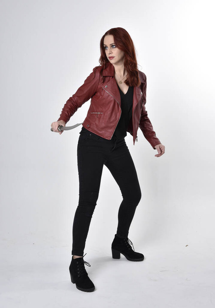 Ritratto di bella ragazza dai capelli rossi con jeans neri e stivali con giacca di pelle. intera lunghezza in piedi posa tenendo coltello uno sfondo di studio
. - Foto, immagini