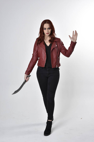 Portrait of a pretty girl with red hair wearing black jeans and boots with leather jacket.  full length standing pose holding knife a studio background. - Photo, Image
