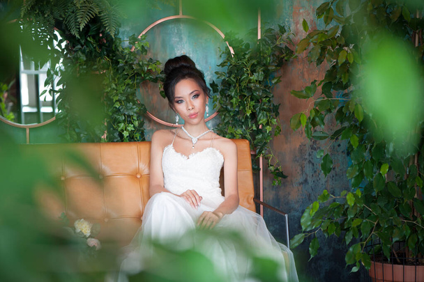 Mariée asiatique avec des fleurs en robe blanche assise sur canapé salle florale intérieure
 - Photo, image