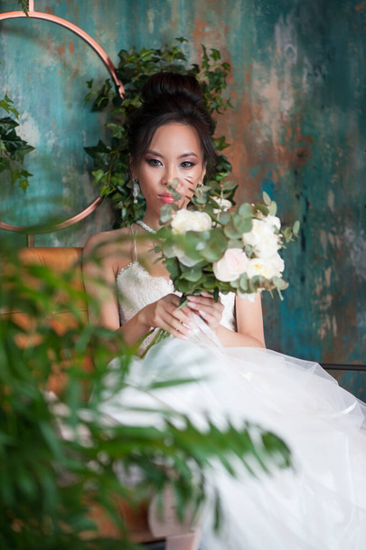 Asian bride with flowers in white dress sitting on sofa indoor floral room - 写真・画像