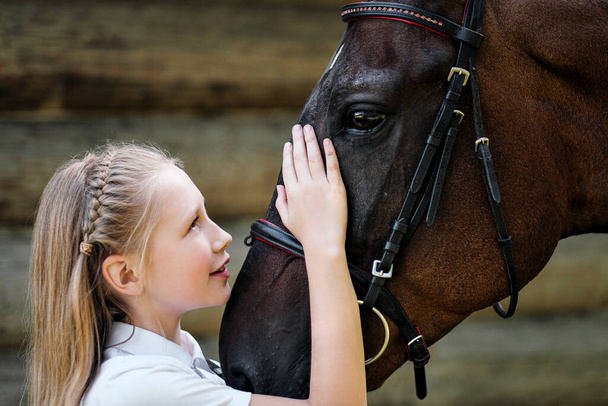 A teenage girl jockey stands next to a brown horse and hugs her. Against the background of a wooden wall stables. - Foto, imagen