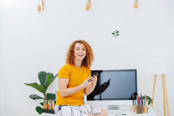 Happy woman artist with gorgeous curly red hair sitting on her work desk with crayons and pc screen. She's holding phone in her hands. With shiny smile on her face. - Photo, Image