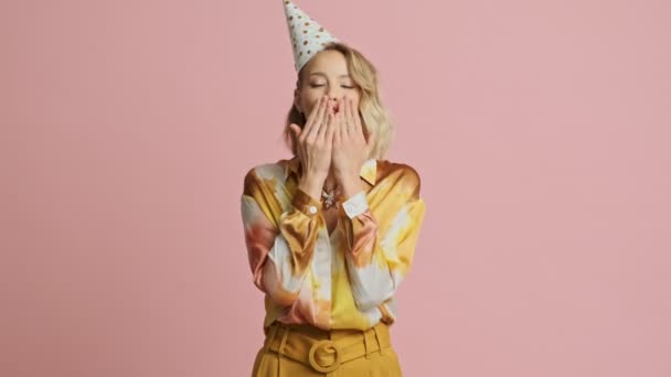 Smiling pretty blonde woman in colorful elegant costume and party cone sending air kiss at the camera over pink background - Imágenes, Vídeo
