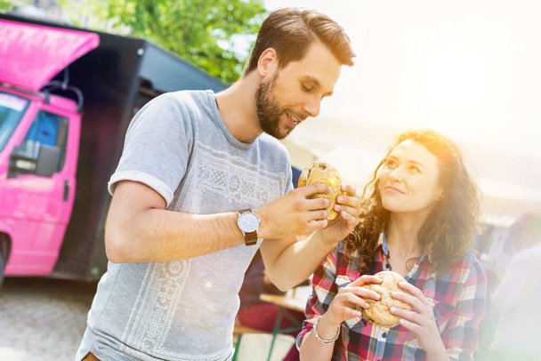 Young attractive couple eating hamburgers against food truck with lens flare in background - Fotoğraf, Görsel