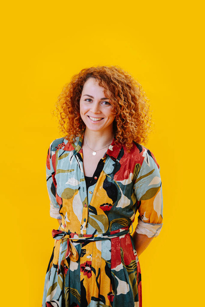 Happy beautiful young woman in a colorful dress with red curly hair and hands behind her back over yellow. Smiling at the camera. - Фото, изображение