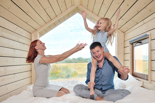 Jovem casal familiar caucasiano com filha bebê em uma pequena casa rústica moderna com uma grande janela. Deitado na cama, abraçando, brincando e olhando pela janela
. - Foto, Imagem