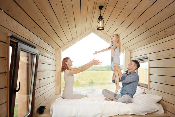 Fiatal kaukázusi házaspár kislányával egy kis modern rusztikus házban, nagy ablakkal. Feküdni az ágyon, ölelkezni, játszani és kinézni az ablakon. - Fotó, kép