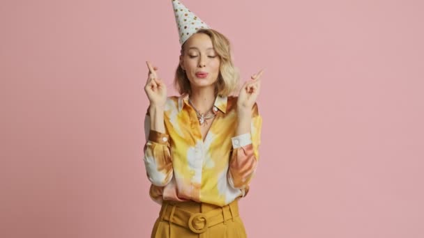 Focused smiling pretty blonde woman in colorful elegant costume and party cone making a wish with crossed fingers gestures over pink background - Felvétel, videó