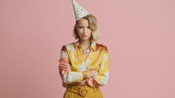 Displeased pretty blonde woman in colorful elegant costume and party cone standing with crossed arms and looking at the camera over pink background - Кадри, відео