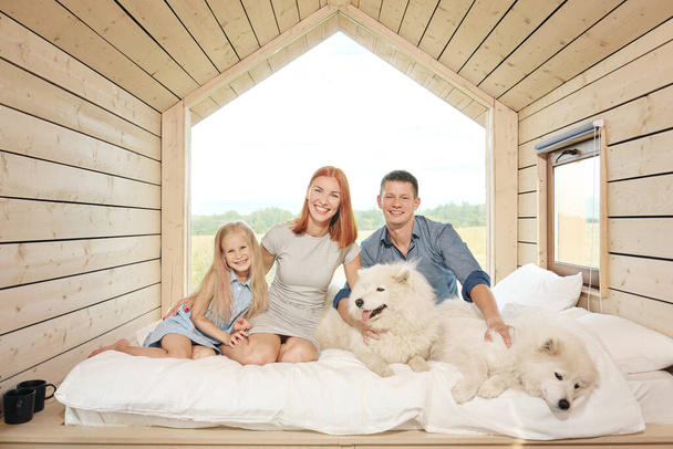 Mladý kavkazský rodinný pár s dcerou v malém moderním venkovském domku s velkým oknem. Ležel na posteli, objímal se, hrál si a díval se z okna. Dva Samoyedi. Víkendová dovolená - Fotografie, Obrázek