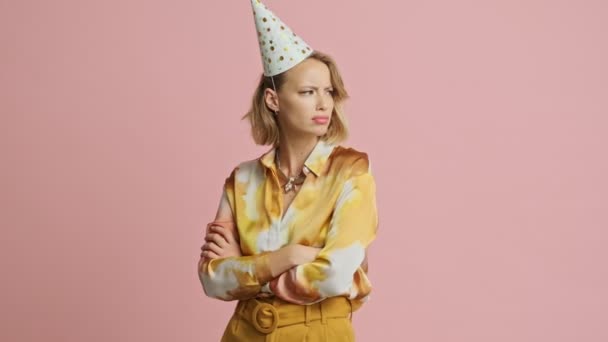 Upset pretty blonde woman in colorful elegant costume and party cone standing with crossed arms and looking around over pink background - Felvétel, videó