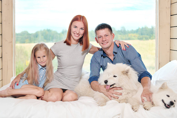 Young Caucasian family couple with baby daughter in a small modern rustic house with a large window. Lying on the bed, hugging, playing and looking out the window. Two Samoyeds. Weekend vacation - Fotografie, Obrázek