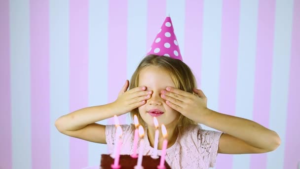 Χαρούμενη έκπληξη, κλείσε τα μάτια κοριτσάκι με το ροζ καπέλο σβήνοντας κεριά σε μια τούρτα γενεθλίων - Πλάνα, βίντεο