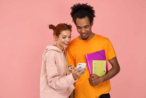 Positive young people focused on a smartphone, watch funny video online, dressed in casual clothes, against a pink background. Happy black guy with notepads looks at ginger girlfriends gadget - Foto, Imagem