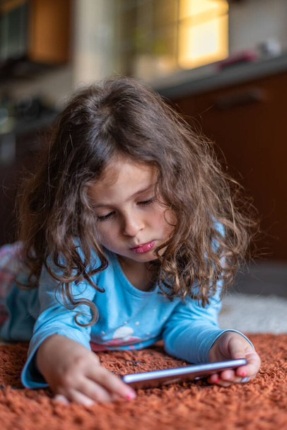 自宅のリビングルームでデジタルスマートフォンの携帯電話を見て横になってかわいい幸せな女の子。家族活動のコンセプト. - 写真・画像