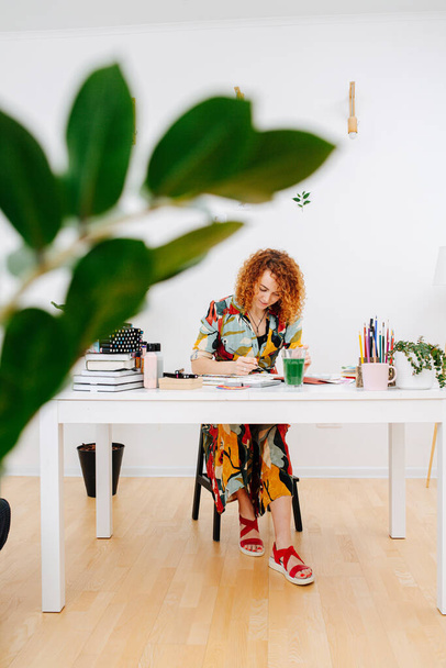Longshot of a focused ginger artist woman in colorful dress painting behind work desk with a brush. Shot through a plant branch. - Фото, зображення