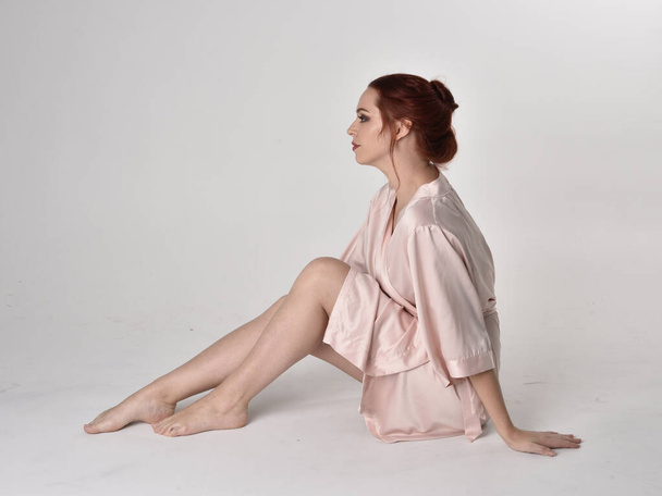 Portrait d'une jolie fille aux cheveux roux portant une robe de soie rose. pose assise sur un fond de studio
. - Photo, image