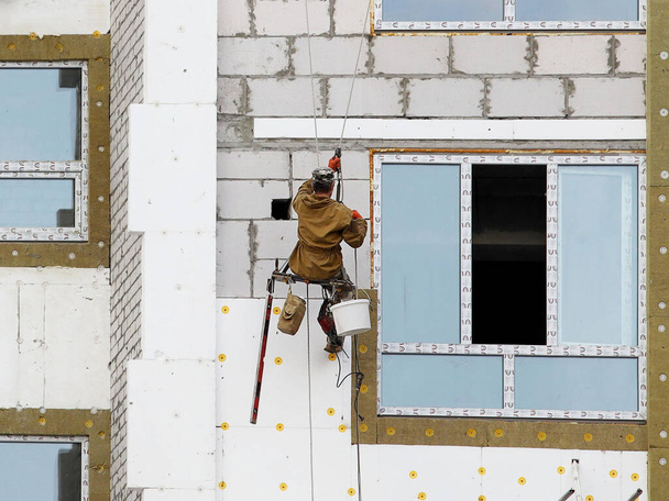 A builder in climbing equipment insulates the facade of a high-rise building with foam plastic. Safety measures for high-altitude construction work. Heat conservation in homes and energy savings. - Photo, Image