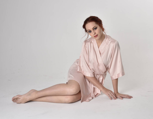 Portrait d'une jolie fille aux cheveux roux portant une robe de soie rose. pose assise sur un fond de studio
. - Photo, image