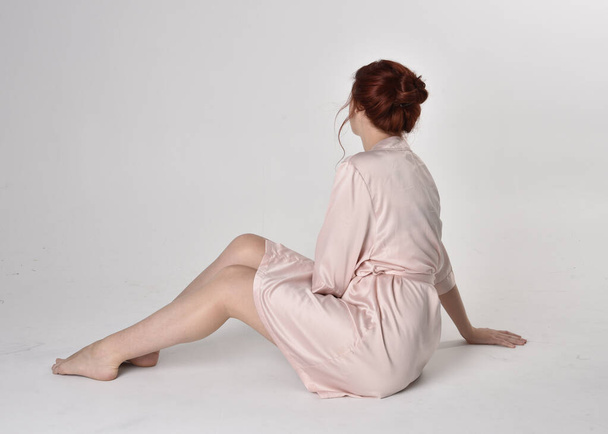 Porträt eines hübschen Mädchens mit roten Haaren in einer rosafarbenen Seidenrobe. sitzende Pose auf einem Studiohintergrund. - Foto, Bild