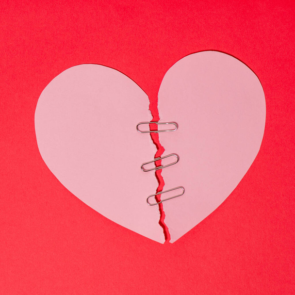 Red paper heart torn in half secured with safety clip on red background - 写真・画像