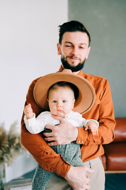 un giovane padre alla moda in camicia arancione di tendenza gioca con il suo giovane figlio in un cappello marrone tra le braccia in uno spazioso monolocale elegante
. - Foto, immagini